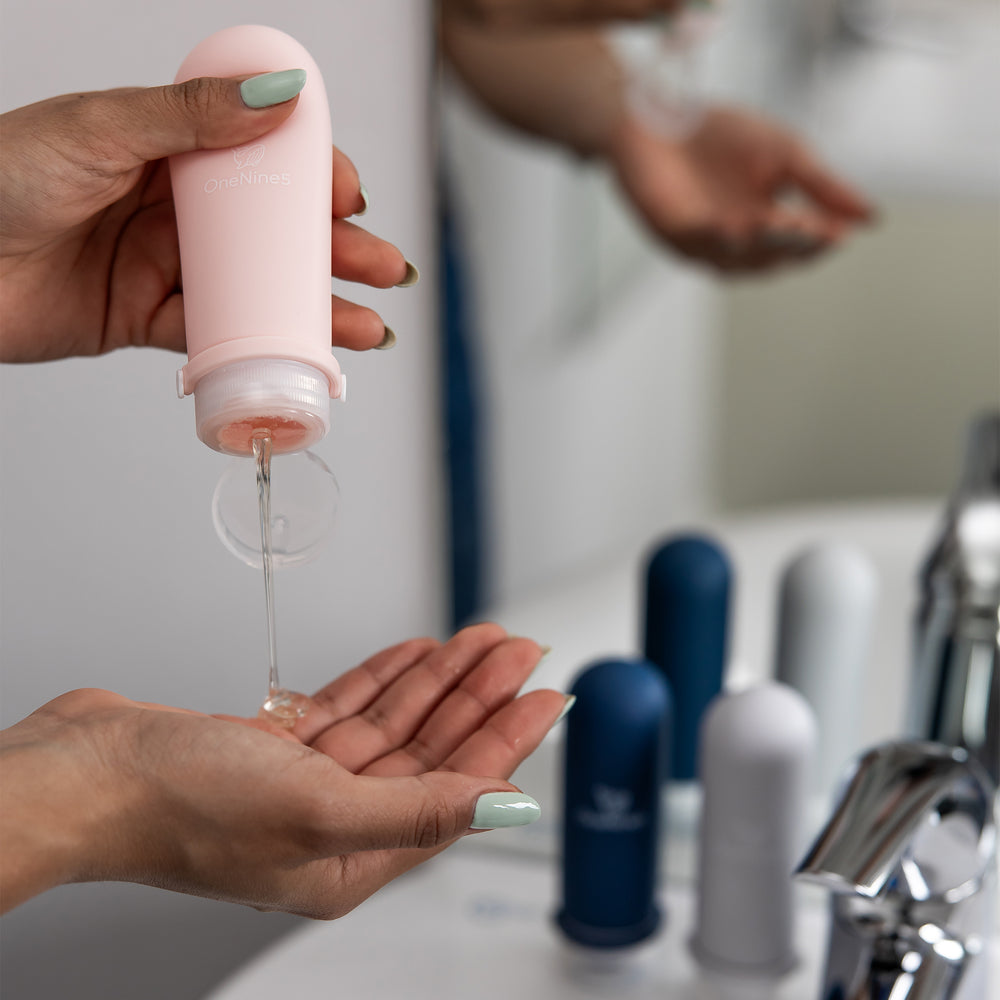 A female in a bathroom, squeezing the soft silicone OneNine5 pink travel bottle into her hand, over the sink. With the leakproof cap (lid) flipped open.