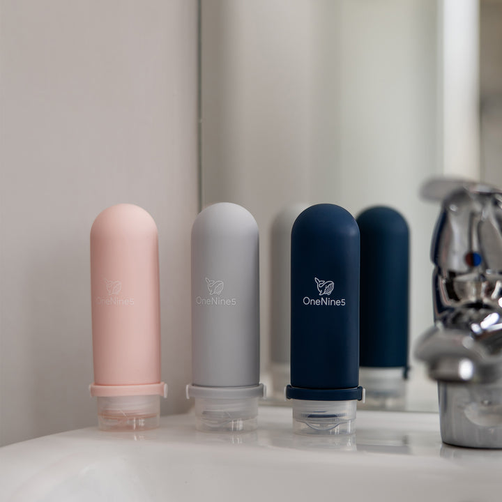3-pack of mixed colour OneNine5 travel bottles on a white bathroom sink next to a chrome tap.