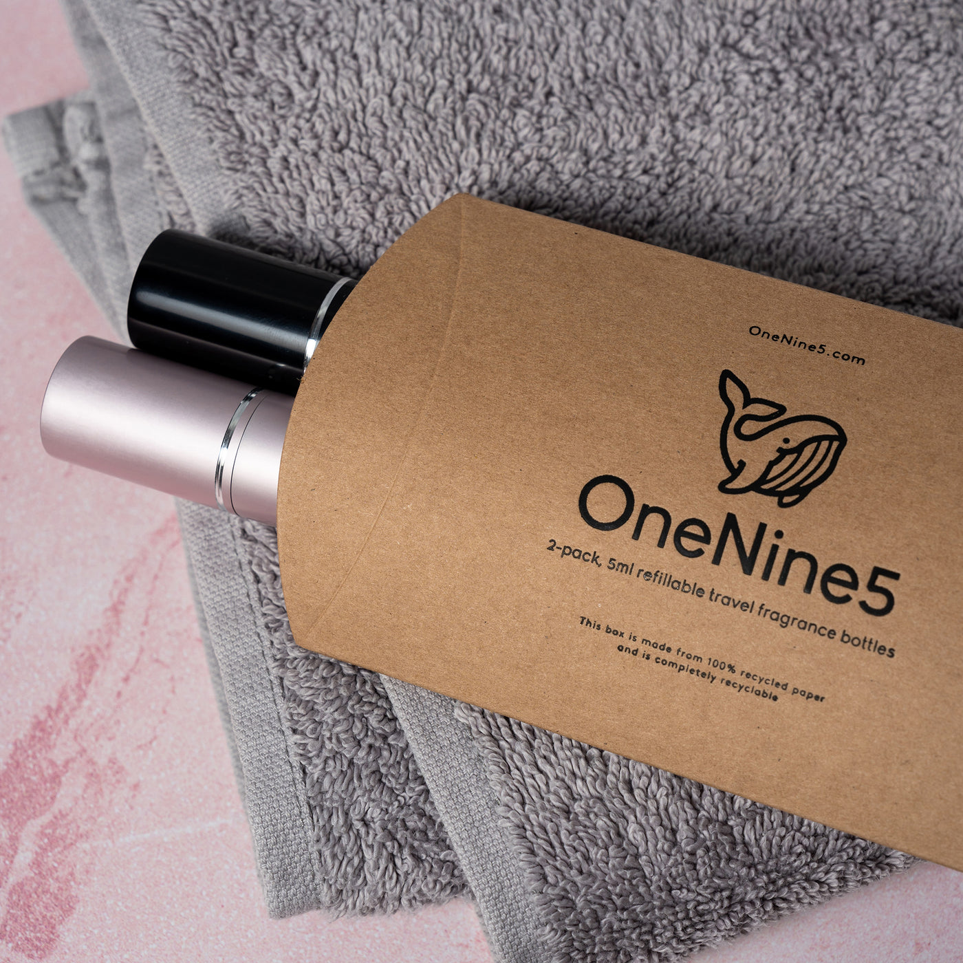 A pink and black OneNine5 fragrance bottle, poking out of the natural kraft paper packaging, resting on a towel in the bathroom