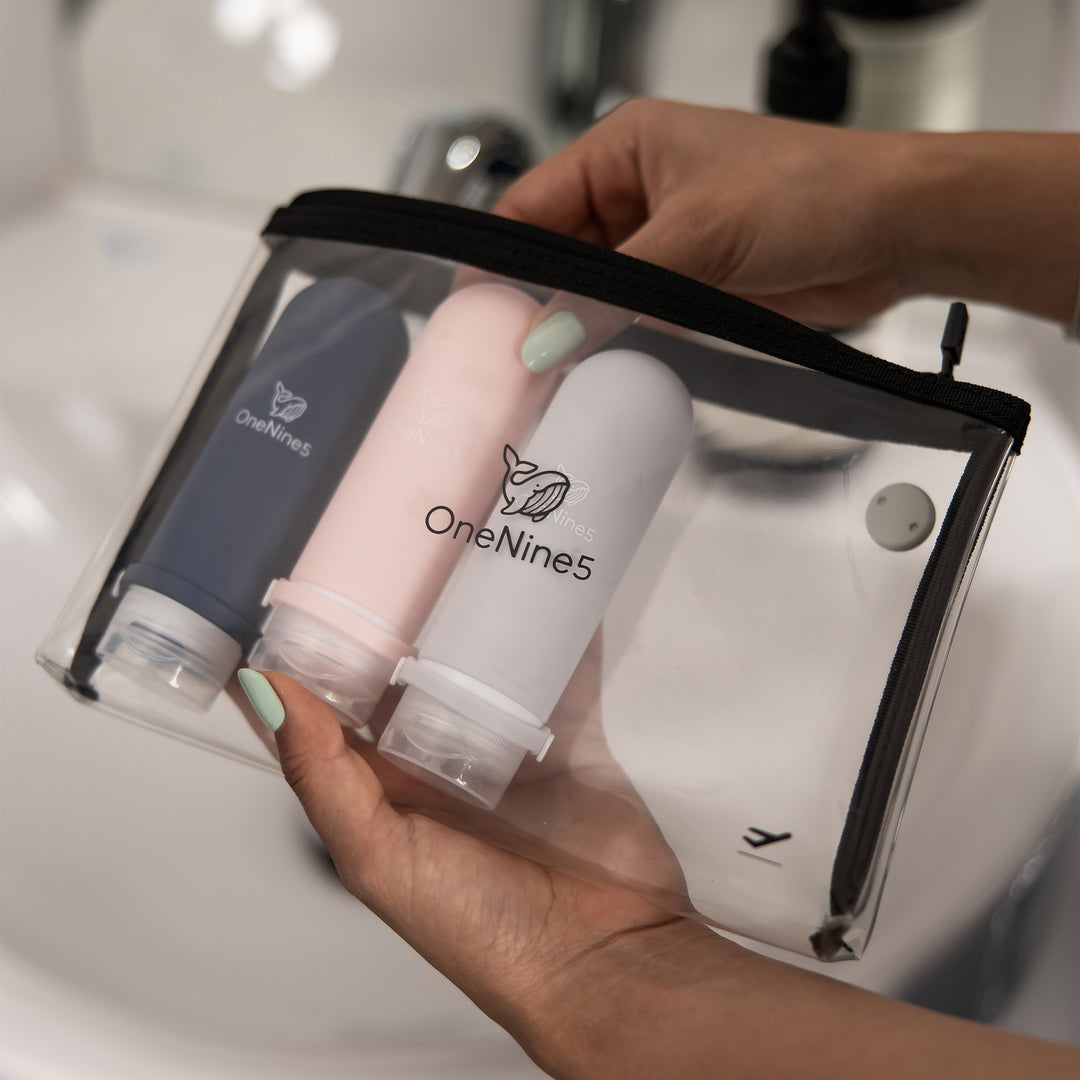 A woman, in the bathroom, packing a blue, pink and grey silicone bottles inside the removable, clear OneNine5 toiletry/ cosmetic bag.