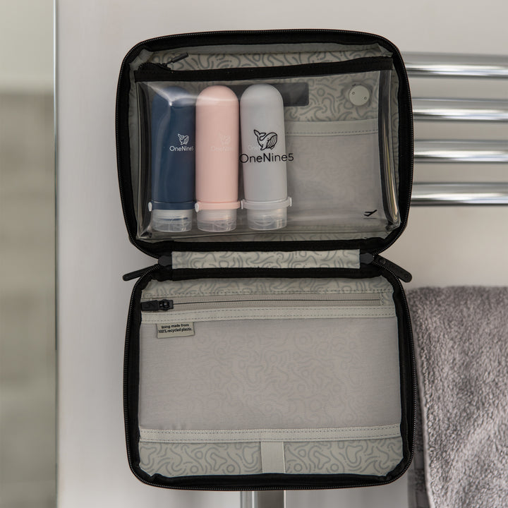 A blue, pink, and grey silicone travel bottle inside the clear toiletry pouch. The pouch is attached inside the OneNine5, Miho Black wash bag, using the hanging hook, in the bathroom.