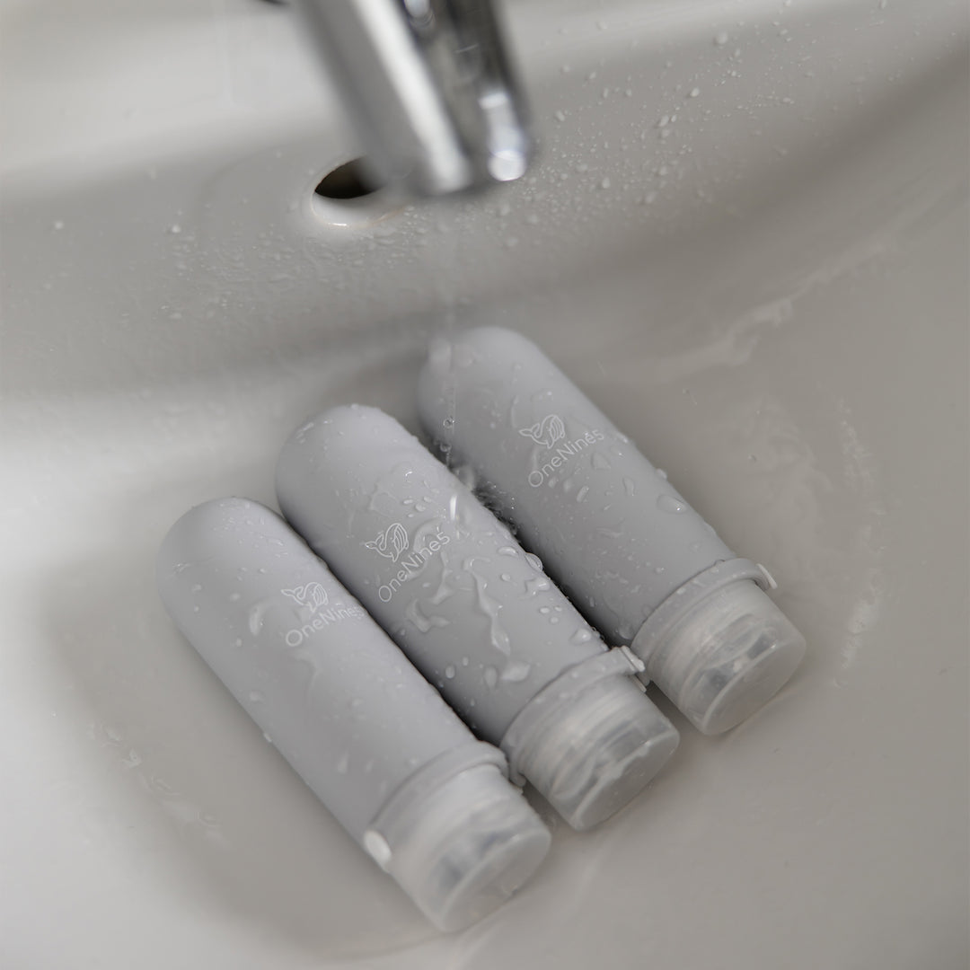 Three grey silicone travel bottles in the bathroom sink. A white OneNine5 logo is visible on the reusable bottles, and they are being splashed with water.