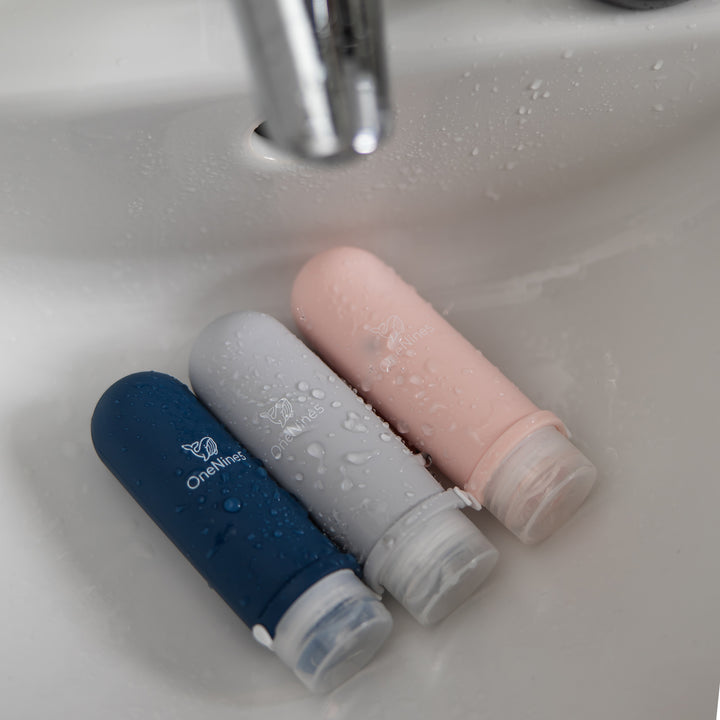 A blue, pink, and grey silicone travel bottle in the bathroom sink. A white OneNine5 logo is visible on the reusable bottles, and they are being splashed with water from the tap.