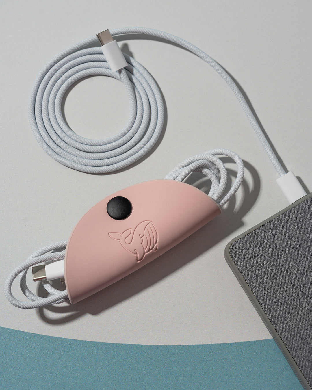 A pink OneNine5 cable tidy, with a 1 metre Apple USB-C wire coiled inside. Next to this is a grey power bank and a coiled white charging cable, on a a white background.