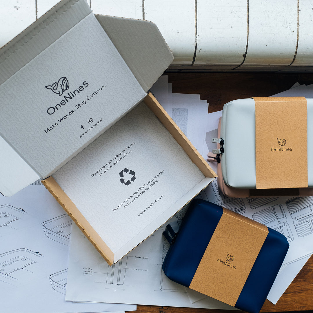 Birds-eye view of a Havelock Blue and Moeraki Grey Eco-Conscious Wash Bags on a desk. With an open, branded shipping box and technical drawings of the wash bag