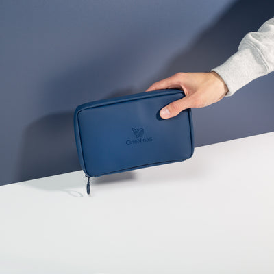 A male hand coming into shot from the right side to hold the Havelock Blue Eco Essentials Pouch in front of a blue wall. The bag is zipped closed and the debossed OneNine5 is visible on the front of the pouch.