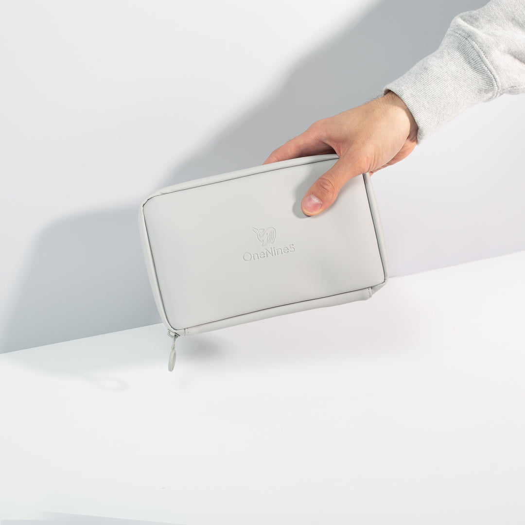 A male hand coming into shot from the right side to hold the Moeraki Grey Eco Essentials Pouch in front of a grey wall. The bag is zipped closed and the debossed OneNine5 is visible on the front of the pouch.