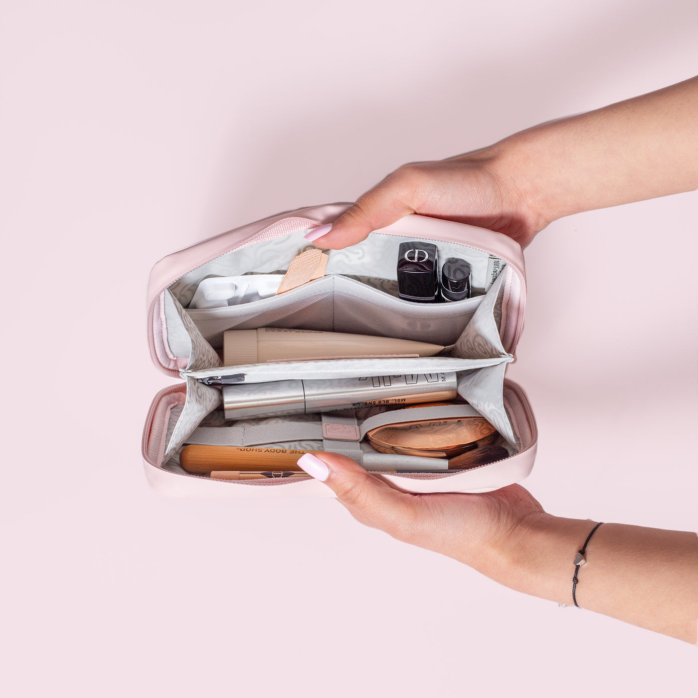 Female hands coming into shot from the right side holding open the Komodo Pink Eco Essentials Pouch to provide an internal view of the storage. On a pink background, packed inside the mesh pockets and elastic loops are painkillers, two fabric plasters, Christian Dior lipstick, Aesop lipbalm, Le Labo hand cream, Milk Mascara, Charlotte Tilbury face powder, Body Shop vegan makeup brush and a Charlotte Tilbury eyebrow pencil.