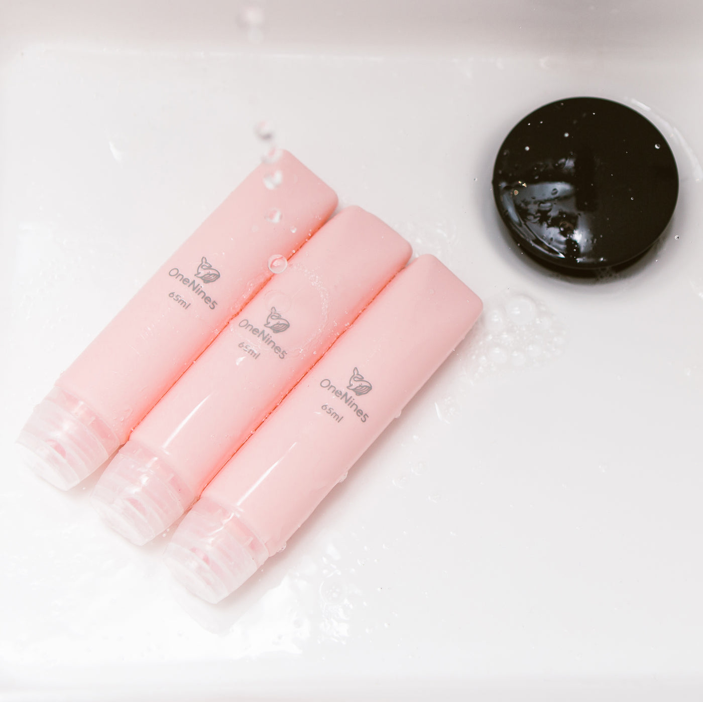 three pink silicone travel bottles in the bathroom sink. A grey OneNine5 logo is visible on the reusable bottles and they are being splashed with water