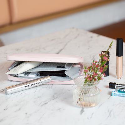 The Komodo Pink Eco Essentials Pouch is laying flat on a marble table. The pouch is zipped open with Le Labo hand cream, painkillers and Milk Mascara inside. On the table, next to the pouch is a small small glass vase with flowers in, a vegan and reusable lipsstick, NARS concealer, Wrigleys chewing gum and Aesop lip balm.