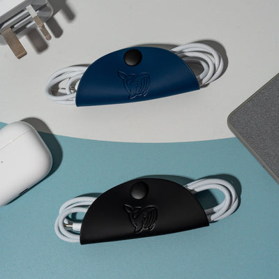 A black and blue OneNine5 cable tidy, with a 1 metre Apple USB-C wire inside. The Cable Tacos are surrounded by Apple Airpods, USB-C plug and grey power bank.