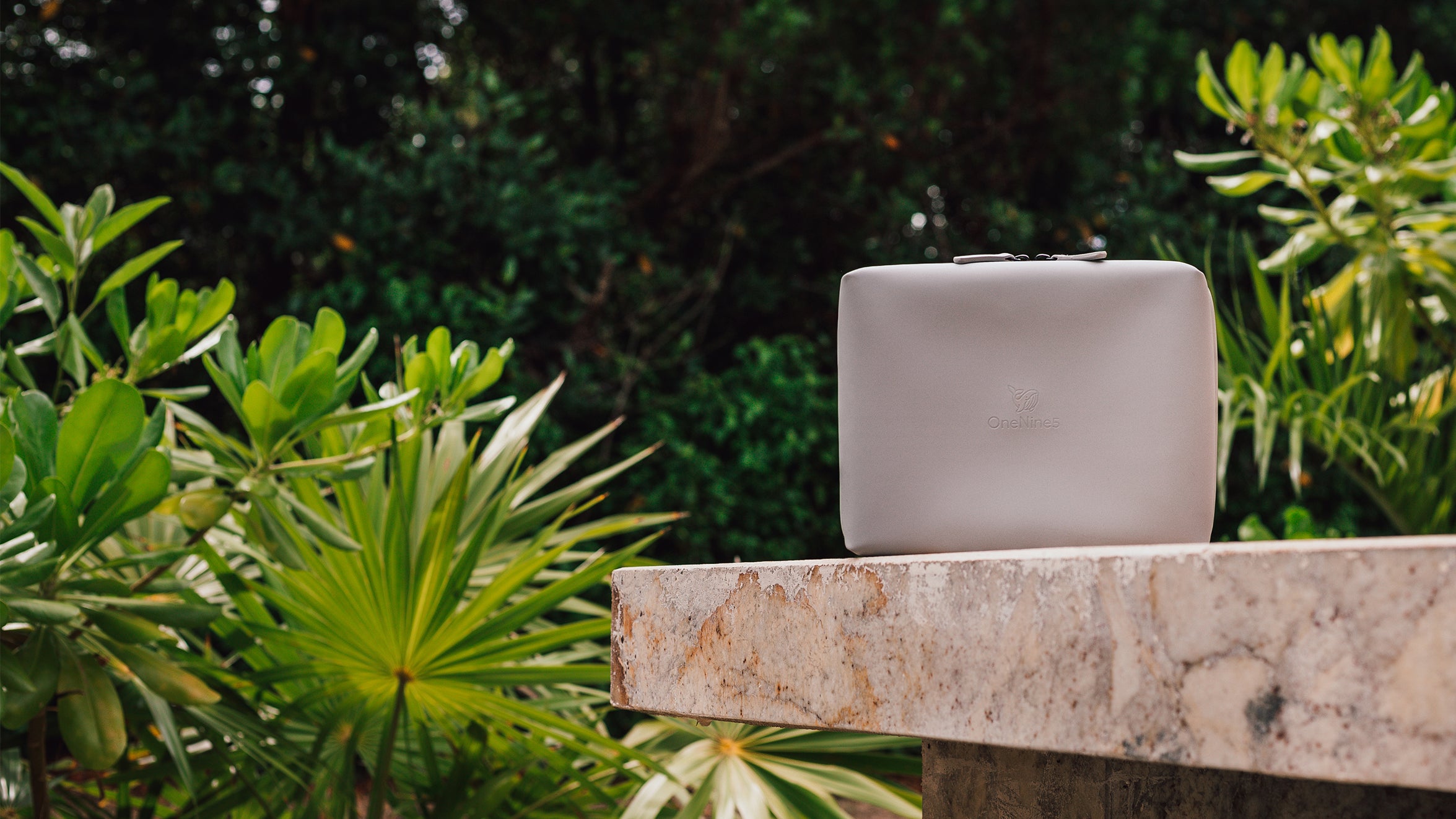 The OneNine5 Moeraki Grey Wash Bag in the side of a swimming pool with green foliage in the background