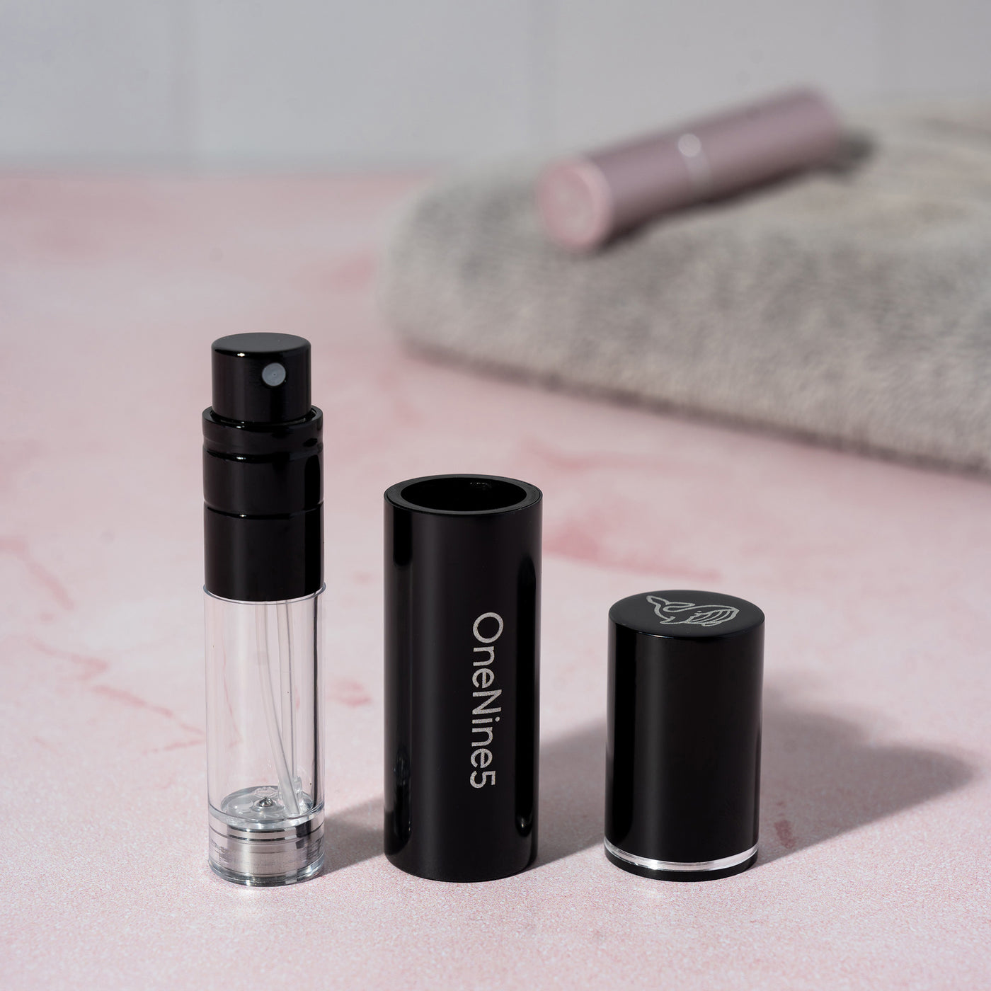 The 3 components of the black OneNine5 refillable fragrance bottle - the images shows the pump, case and magnetic lid in line. A pink bottle and towel is blurred in the background