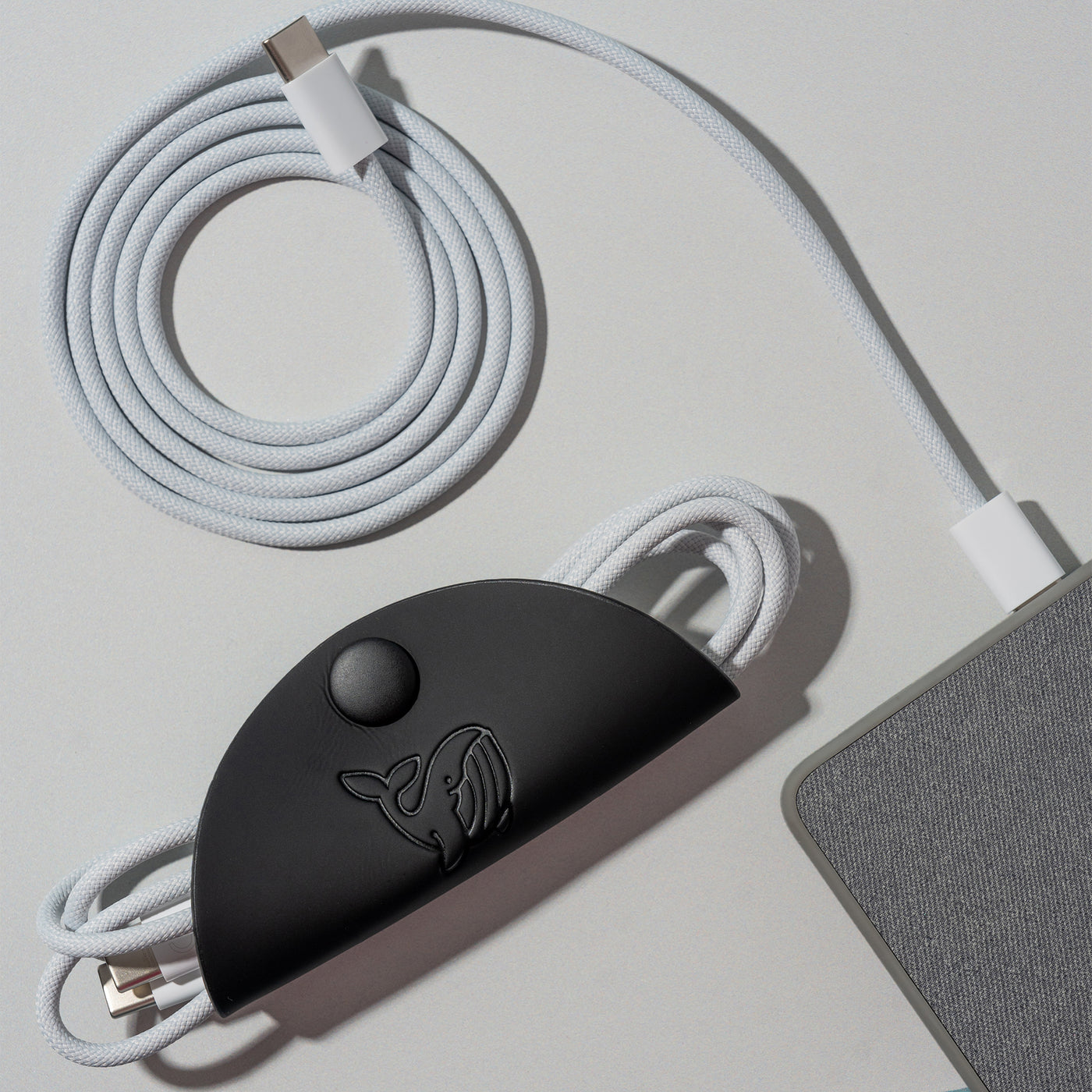 A black OneNine5 cable tidy, with a 1 metre Apple USB-C wire coiled inside. Next to this is a grey power bank and a coiled white charging cable, on a a white background.