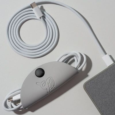 A grey OneNine5 cable tidy, with a 1 metre Apple USB-C wire coiled inside. Next to this is a grey power bank and a coiled white charging cable, on a a white background.