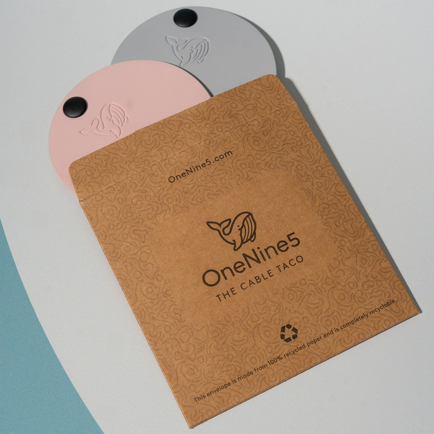 A grey and pink OneNine5 cable tidy, poking out of the unbleached and plastic-free kraft paper packaging.