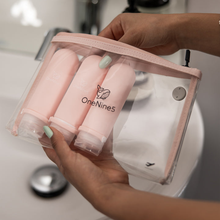 A woman, in the bathroom, packing 3 pink silicone bottles inside the removable, clear OneNine5 toiletry/ cosmetic bag.