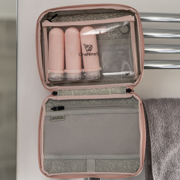 Three pink silicone travel bottles in the bathroom sink. A white OneNine5 logo is visible on the reusable bottles, and they are being splashed with water.