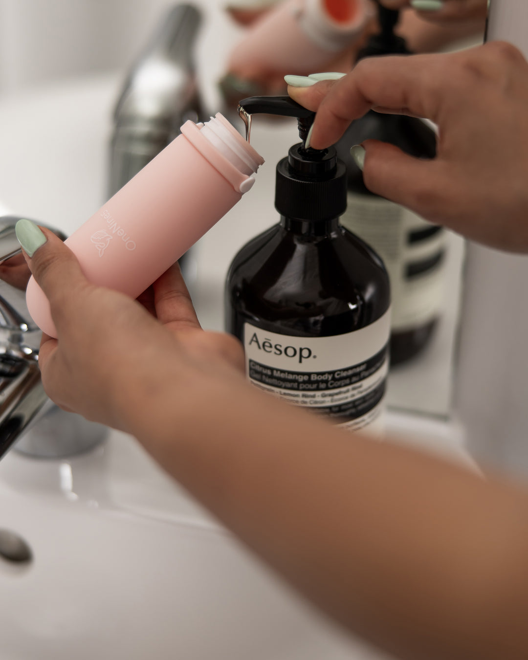 A female refilling the pink OneNine5 silicone travel bottle with a large bottle of Aesop body wash bag in the bathroom