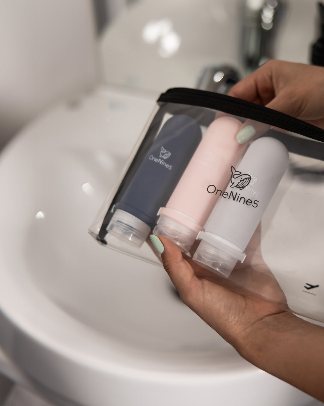 A female in the bathroom. Packing a blue, pink and grey OneNine5 refillable bottle inside a clear toiletry pouch