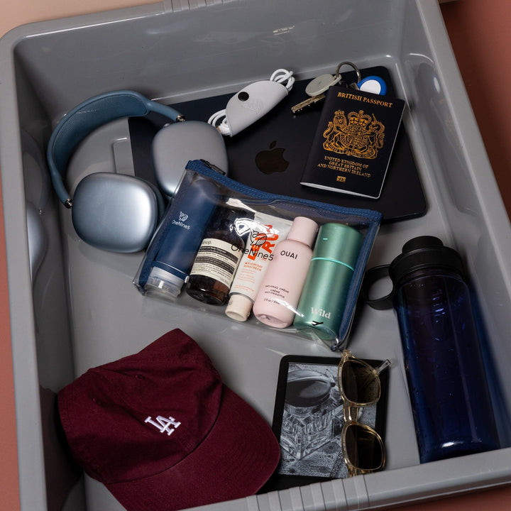 Blue clear liquid travel bag with toiletries packed inside airport security tray alongside passport, headphones and travel essentials