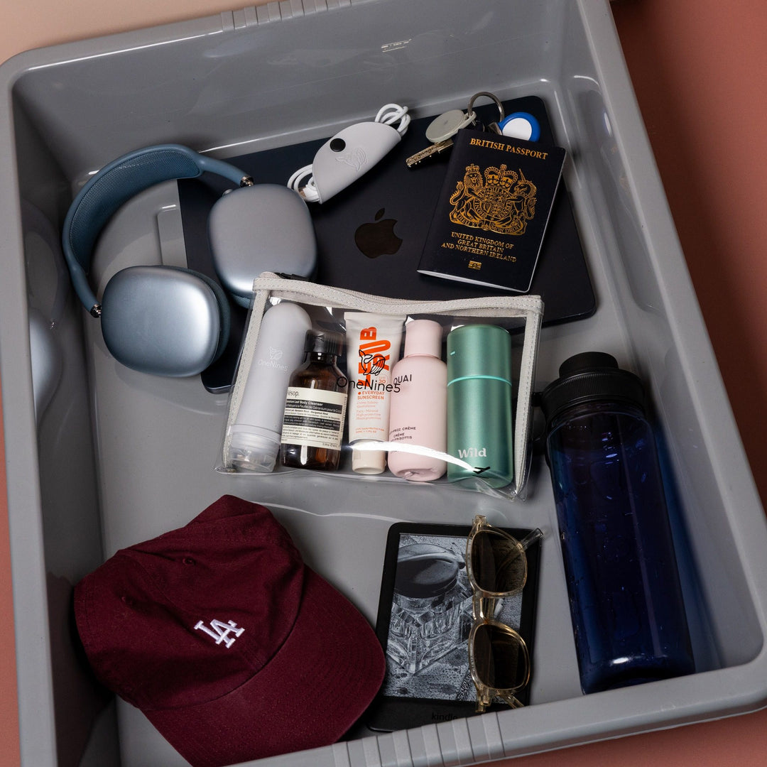 Grey clear liquid travel bag with toiletries packed inside airport security tray with passport, headphones and travel essentials