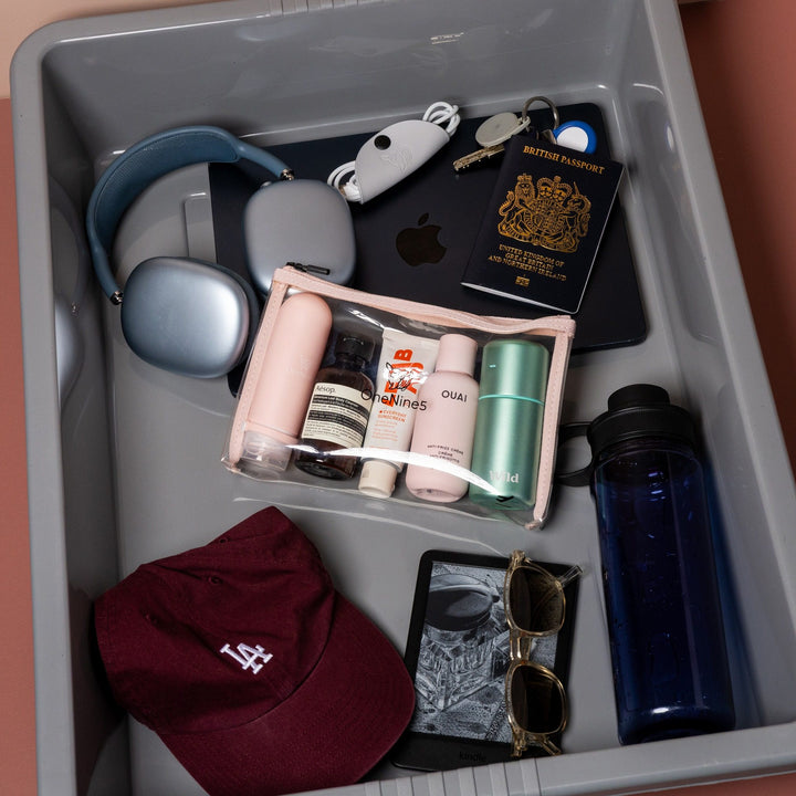 Pink clear liquid travel bag with toiletries inside airport security tray alongside passport, Kindle and travel accessories