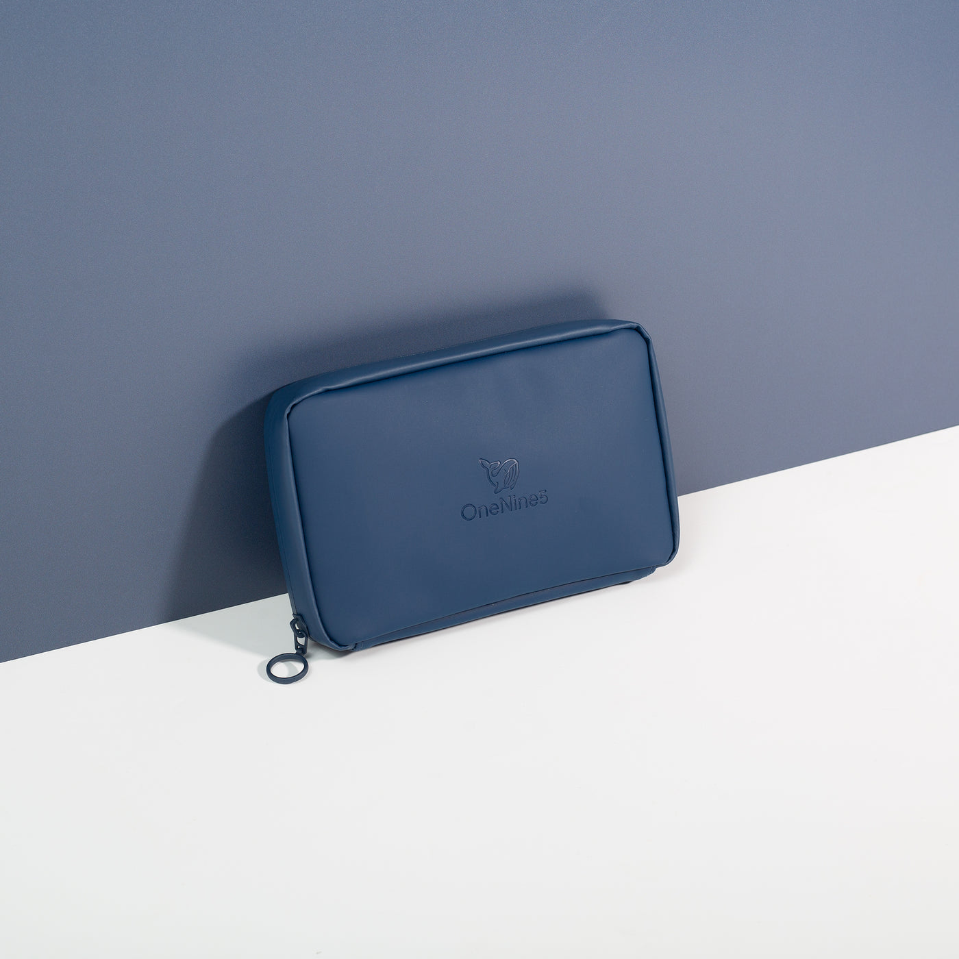 Havelock Blue Eco Essentials Pouch zipped closed and propped up against a blue wall. The debossed OneNine5 Whale logo and metal O-ring zip are visible.
