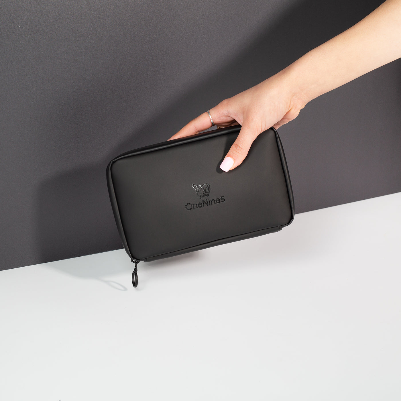 A female hand coming into shot from the right side to hold the Miho Black Eco Essentials Pouch in front of a black wall. The bag is zipped closed and the debossed OneNine5 is visible on the front of the pouch.