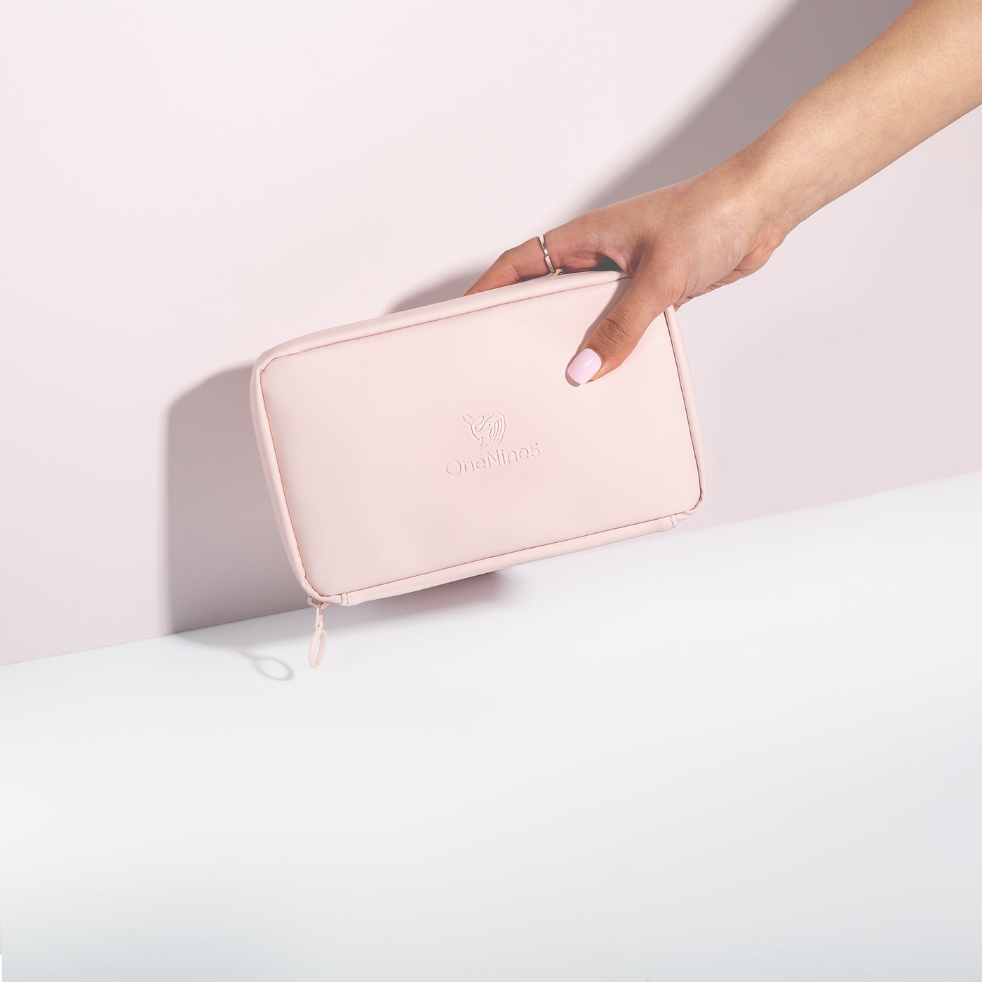 A female hand coming into shot from the right side to hold the Komodo Pink Eco Essentials Pouch in front of a pink wall. The bag is zipped closed and the debossed OneNine5 is visible on the front of the pouch.