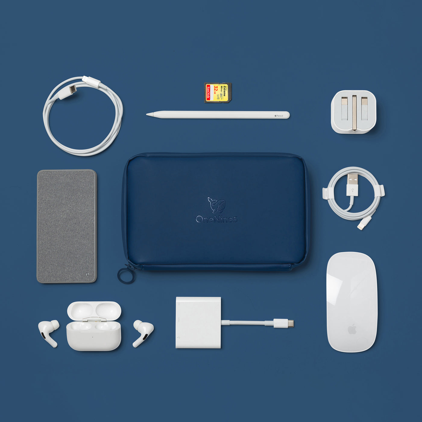 Birdseye image of the Havelock Blue Eco Essentials Pouch on a blue background. the Eco Essentials Pouch is surrounded by a Mophie powerstation mini universal battery, Apple lightning to USB-C cable, Apple pencil, SD card, Apple USB to lightning cable, Apple Mouse, Apple Airpods, USB Power Adapter and Apple USB-C Digital AV Multiport Adapter. These items fit inside the internal pockets and elastic loops.