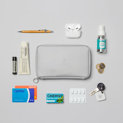 Birdseye image of the Moeraki Grey Eco Essentials Pouch laid on a grey background. the Eco Essentials Pouch is surrounded by Apple Airpods, Beauty Kitchen Organic Vegan Hand Sanitiser, three pound coins, a key with a Tile Bluetooth Tracker attached, painkillers, Chewsy chewing gum, an Oyster card, a Monzo bank card, Le Labo hand moisturiser, Aesop lip balm and a yellow pencil.  These items would fit inside the internal pockets and elastic loops.