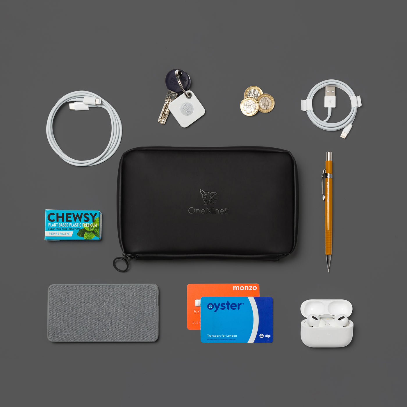 Birdseye image of the Miho Black Eco Essentials Pouch on a black background. the Eco Essentials Pouch is surrounded by an Apple lightning to USB cable, a yellow pencil, Apple Airpods, an Oyster card, Monzo Bank card, Mophie powerstation mini universal battery, Chewsy chewing gum, Apple USB-C to lightning cable, A key attached to a Tile Bluetooth tracker and three pound coins. These items fit inside the internal pockets and elastic loops.