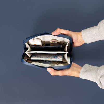 Male hands coming into shot from the right side holding open the Havelock Blue Eco Essentials Pouch to provide an internal view of the storage. On a blue background, packed inside the mesh pockets and elastic loops are La Roche Posay SPF, rolled up Hong Kong Dollars, a small map, a blue UK passport, a Revolut bank card and a suitcase lock.