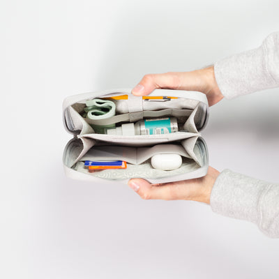 Male hands coming into shot from the right side holding open the Moeraki Grey Eco Essentials Pouch to provide an internal view of the storage. On a grey background, packed inside the mesh pockets and elastic loops are Apple Airpods, an Oyster card, a Monzo bank card, a green face mask, a yellow pencil and Beauty Kitchen Organic Vegan Hand Sanitiser.