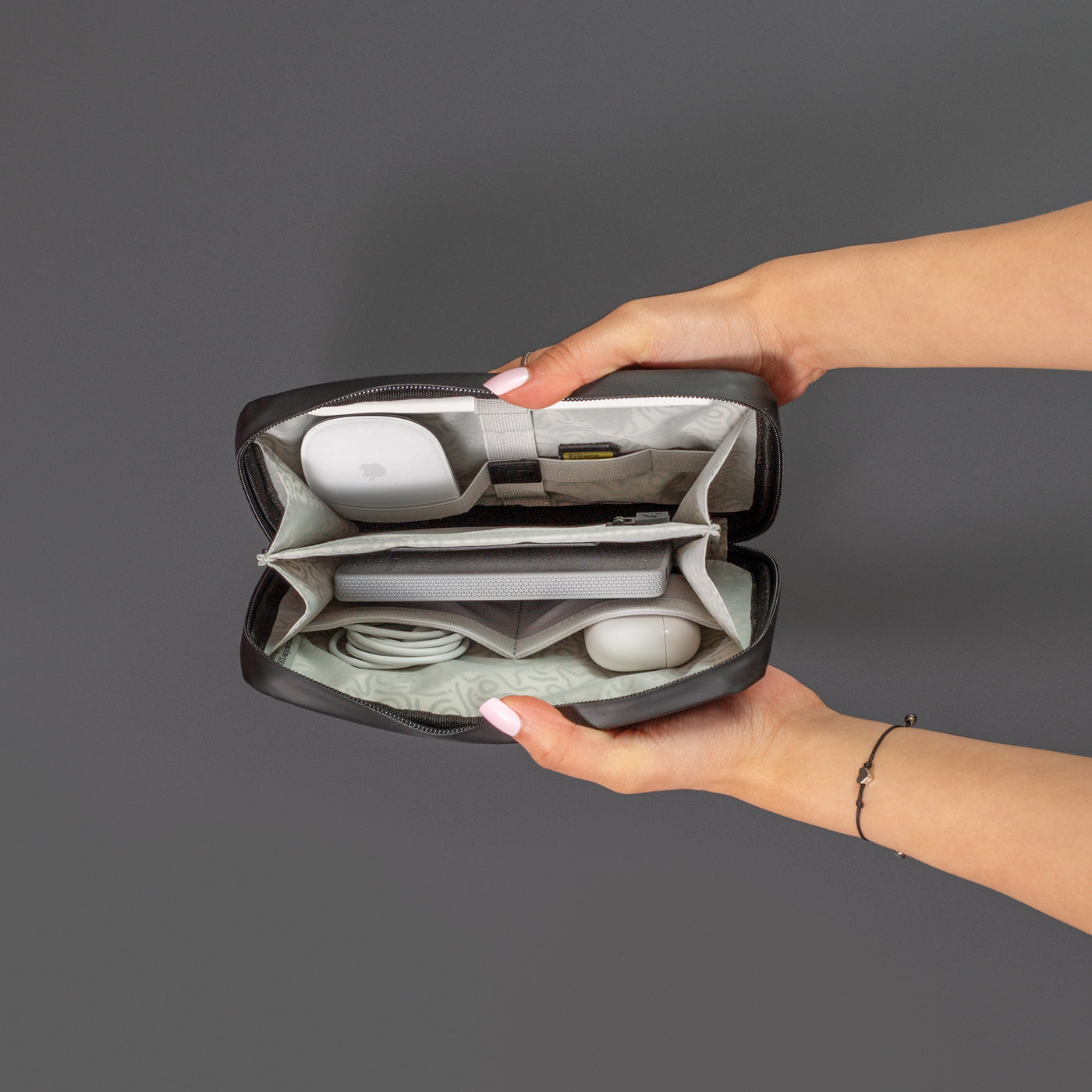 Female hands coming into shot from the right side holding open the Miho Black Eco Essentials Pouch to provide an internal view of the storage. On a black background, packed inside the mesh pockets and elastic loops are Apple Airpods, an Apple Mouse, an SD card, an Apple charging cable and a Mophie powerstation mini universal battery.