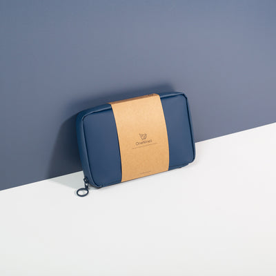 Havelock Blue Eco Essentials Pouch zipped closed and propped up against a blue wall. Packaged in a 100% recycled kraft paper sleeve. The OneNine5 logo is visible on the front of the brown kraft paper sleeve.