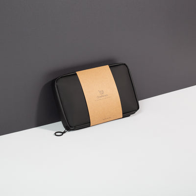 Miho Black Eco Essentials Pouch zipped closed and propped up against a black wall. Packaged in a 100% recycled kraft paper sleeve. The OneNine5 logo is visible on the front of the brown kraft paper sleeve.
