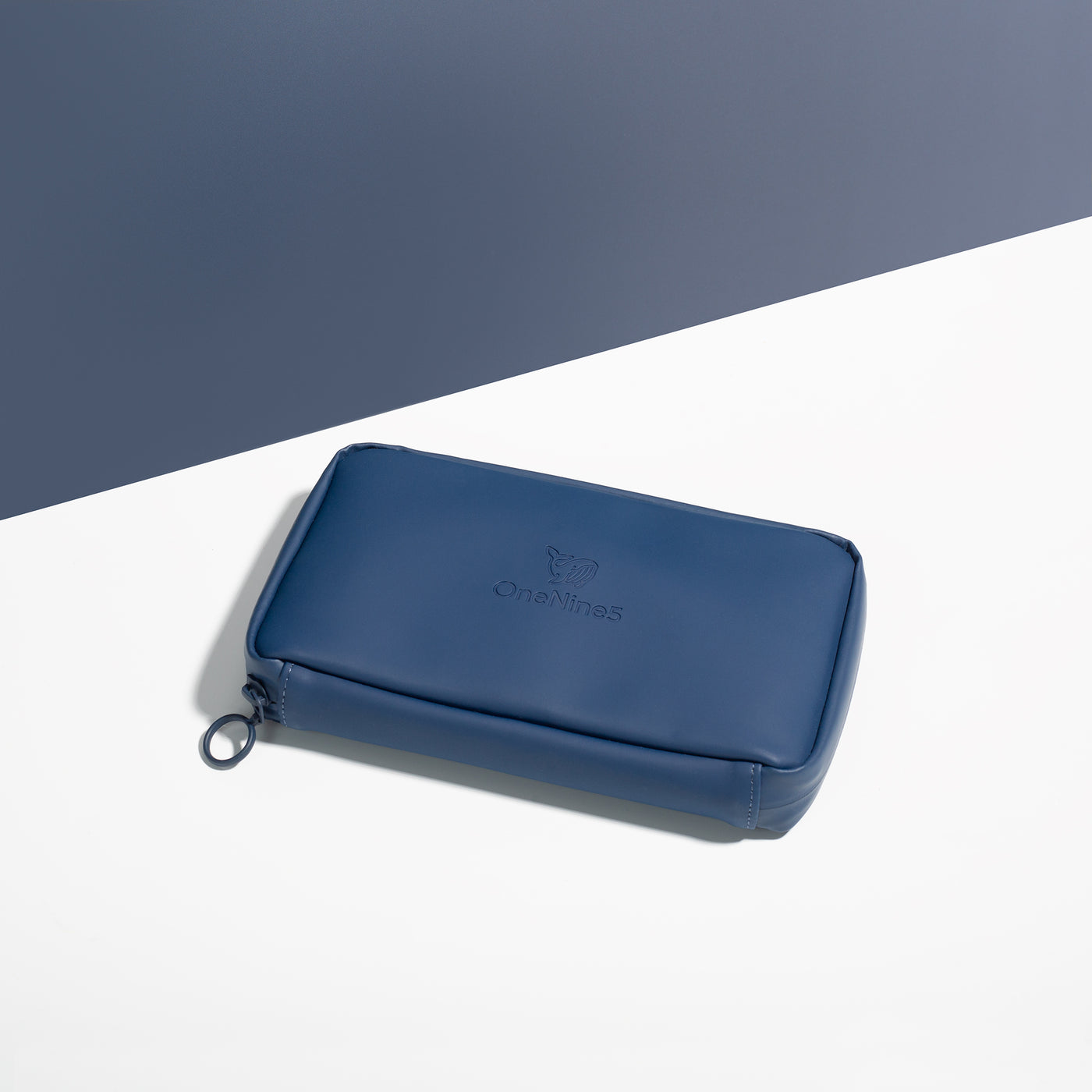 Havelock Blue Eco Essentials Pouch zipped closed and laid flat on a white surface, with a blue background behind. The angle of the image show’s the underside of the pouch. In the foreground, the metal O-ring zipper puller is visible on the left corner of the pouch.