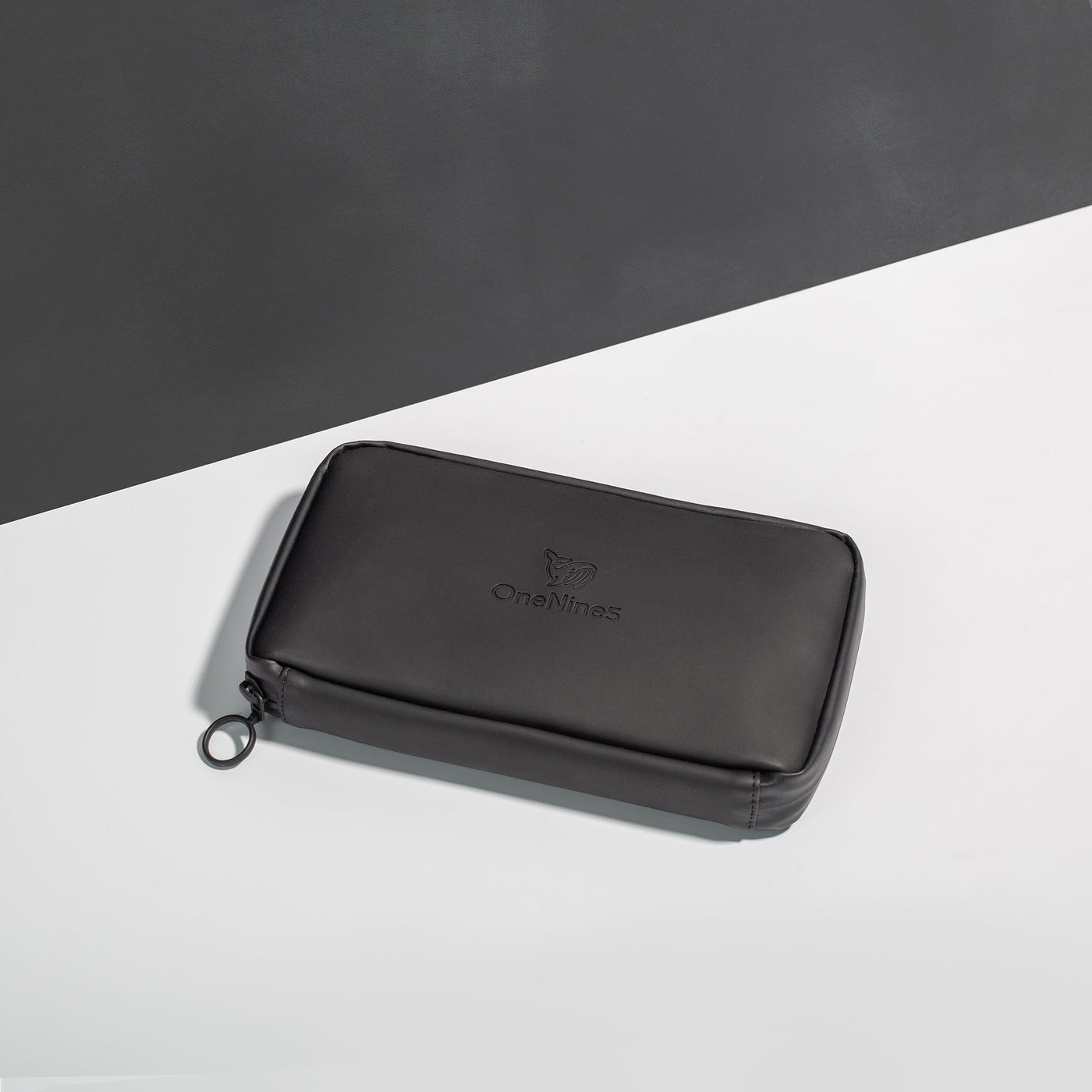 Miho Black Eco Essentials Pouch zipped closed and laid flat on a white surface, with a black background behind. The angle of the image show’s the underside of the pouch. In the foreground, the metal O-ring zipper puller is visible on the left corner of the pouch.