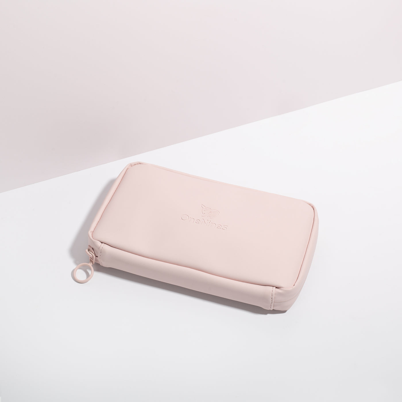 Komodo Pink Eco Essentials Pouch zipped closed and laid flat on a white surface, with a pink background behind. The angle of the image show’s the underside of the pouch. In the foreground, the metal O-ring zipper puller is visible on the left corner of the pouch.