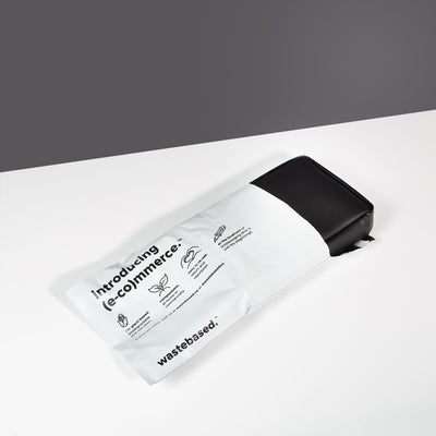 Miho Black Eco Essentials Pouch poking out of a 100% biodegradable and compostable Wastebased shipping bag. The pouch and shipping bag are laid flat on a white surface with a black background.