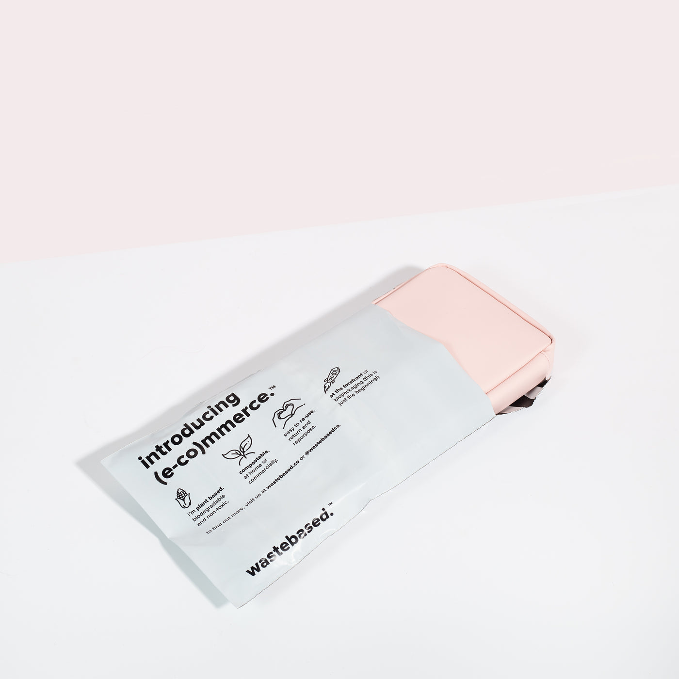 Komodo Pink Eco Essentials Pouch poking out of a 100% biodegradable and compostable Wastebased shipping bag. The pouch and shipping bag are laid flat on a white surface with a pink background.