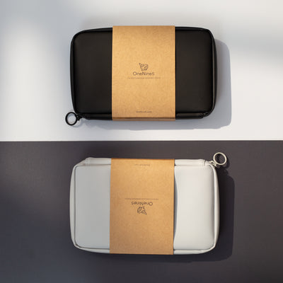 A birds-eye image of the Moeraki Grey Essentials Pouch and Miho Black Essentials Pouch. Each pouch is laid opposite. The grey pouch is on a black background and the black pouch on a grey background. Both Essentials Pouches are packaged in their kraft paper sleeve.