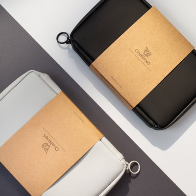 A birds-eye image of the Moeraki Grey Essentials Pouch and Miho Black Essentials Pouch. Each pouch is laid diagonally and opposite each other. The grey pouch is on a black background and the black pouch on a grey background. Both Essentials Pouches are packaged in their kraft paper sleeve.