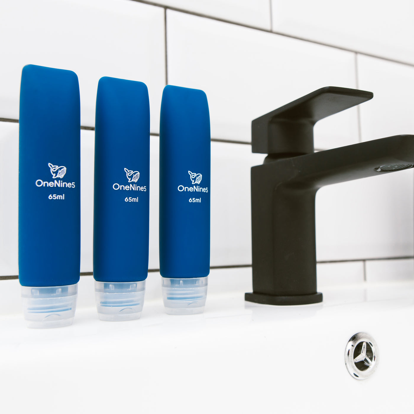 3 pack of OneNine5 blue silicone travel bottles on the bathroom sink, to the right of a black tap