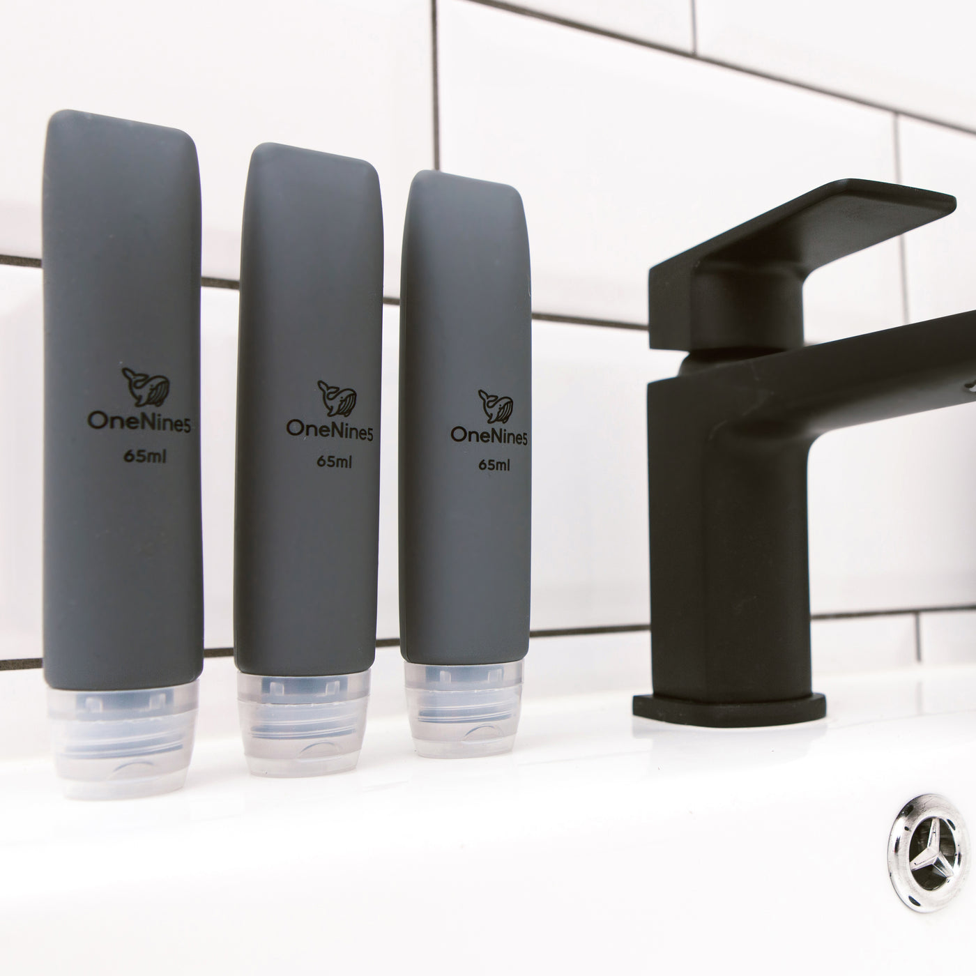 3 pack of OneNine5 grey silicone travel bottles on the bathroom sink, to the right of a black tap