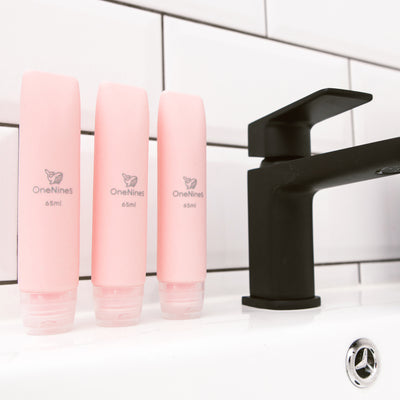 3 pack of OneNine5 pink silicone travel bottles on the bathroom sink, to the right of a black tap