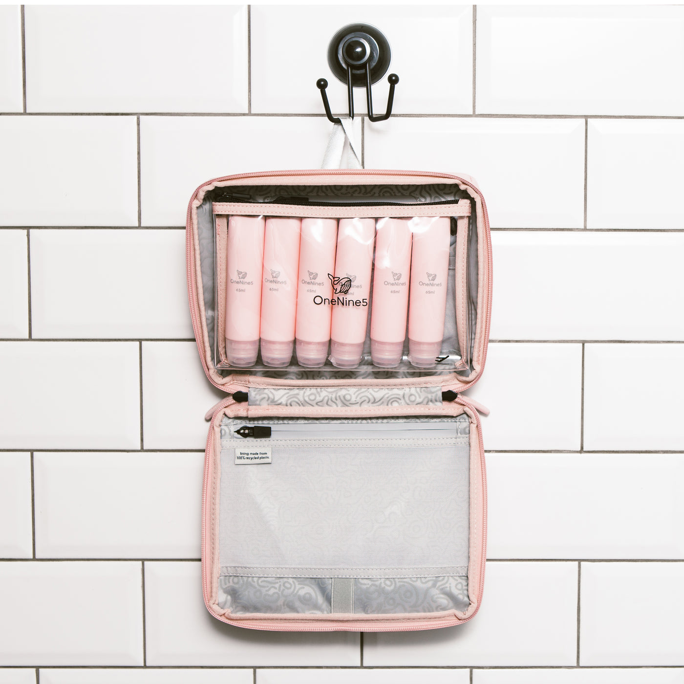 The OneNine5 Komodo Pink, travel wash bag hanging in the bathroom with white tiles. Inside the wash bag is the reusable clear liquid pouch with six pink, reusable silicone bottles inside