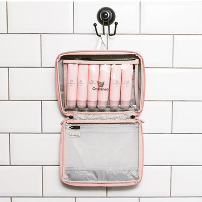 The OneNine5 Komodo Pink, travel wash bag hanging in the bathroom with white tiles. Inside the wash bag is the reusable clear liquid pouch with six pink, reusable silicone bottles inside